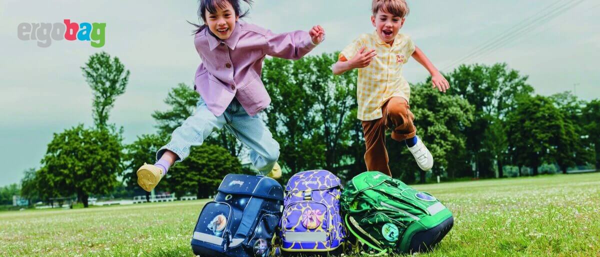 3 Kinder springen auf der Wiese und vor ihnen stehen ihre Ergobag Rucksäcke.
