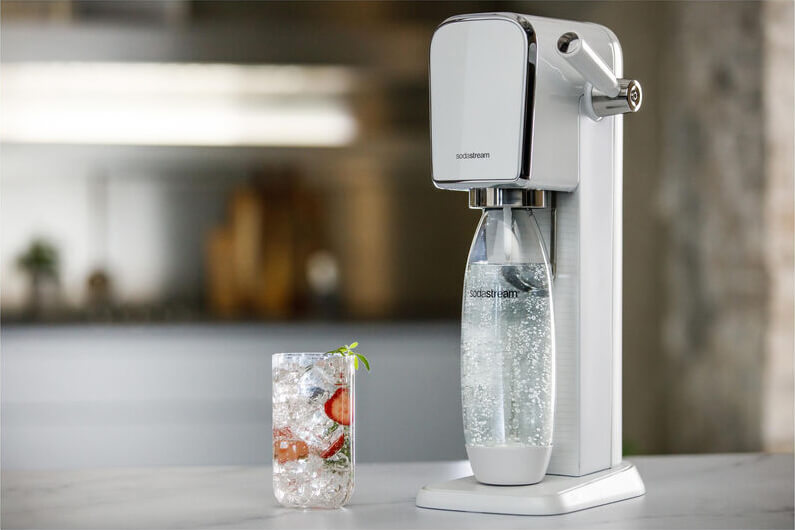 Weisser Sodastream daneben ein Wasserglas mit Früchten